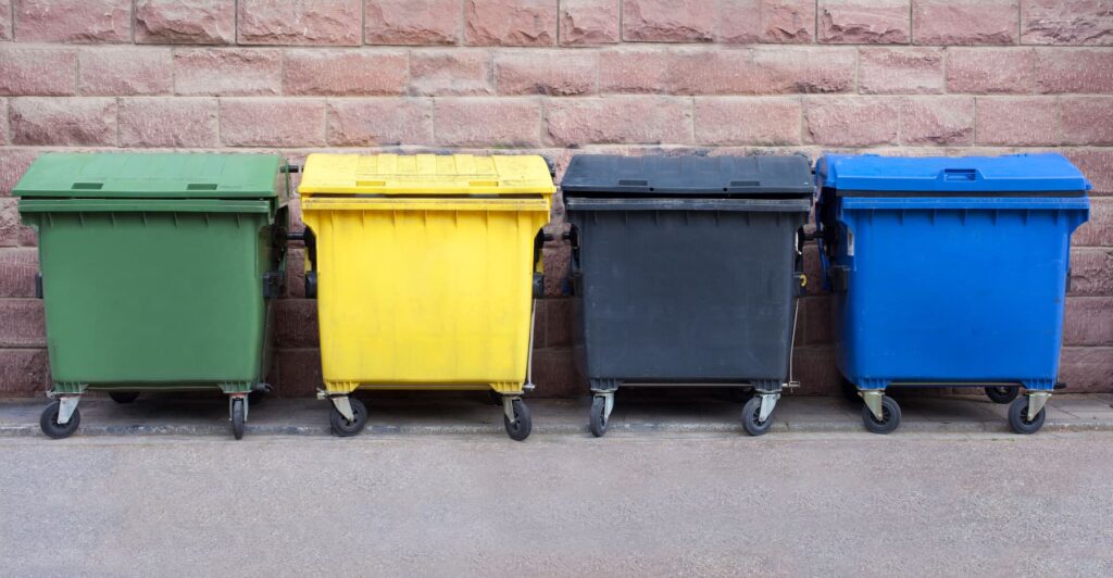 Vier farbcodierte Wertstoffsammler-Container (grün, gelb, schwarz, blau) zur effizienten Mülltrennung an einer Ziegelsteinmauer.