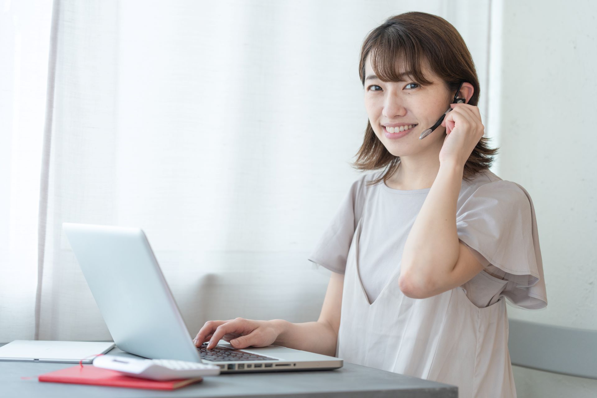 Eine junge Frau mit Headset arbeitet an einem Laptop und lächelt freundlich in die Kamera. Sie verwendet moderne Kommunikationstechnologie, um in einem professionellen Kontext über eine Online-Telefonanlage zu arbeiten.