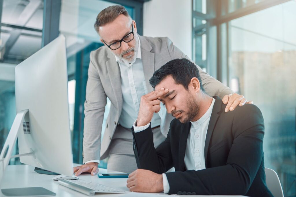 Fuehrungskraft spricht mit gestresstem Mitarbeiter am Schreibtisch – empathische Geste im Buero